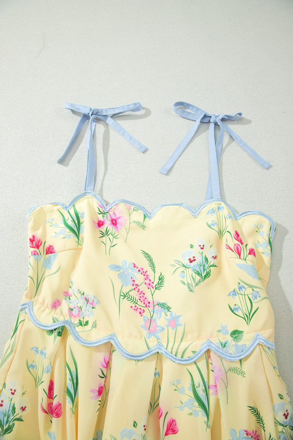 Robe midi crème à fleurs, bretelles nouées bleu clair, haut froncé et bord festonné.